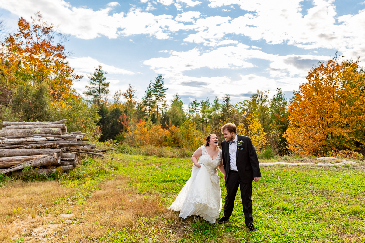 A bride and groom laugh walking together in front of fall foliage and a pile of wood